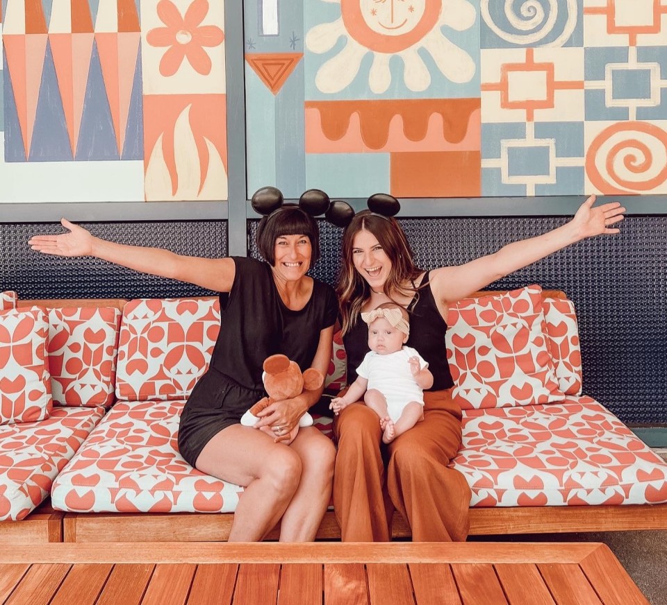 Three generations of women, wearing Mickey Mouse ears, sit on a couch in front of a whimsical mural.