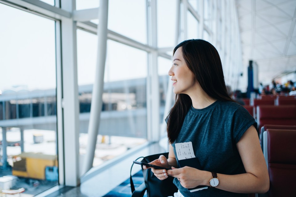 Woman at airport using smartphone, holding passport and boarding pass.