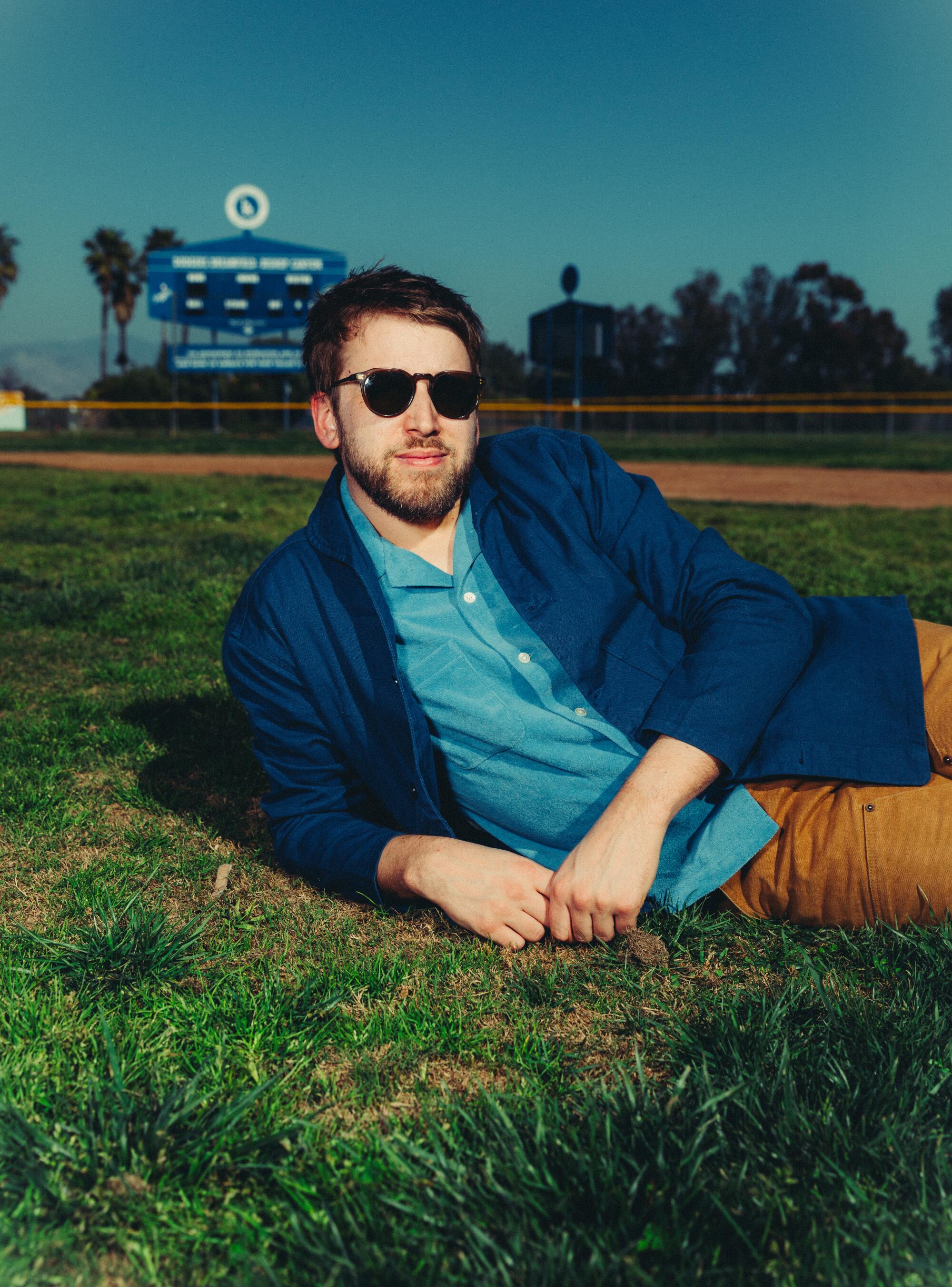 A man in shades lies on the grass of a baseball field.