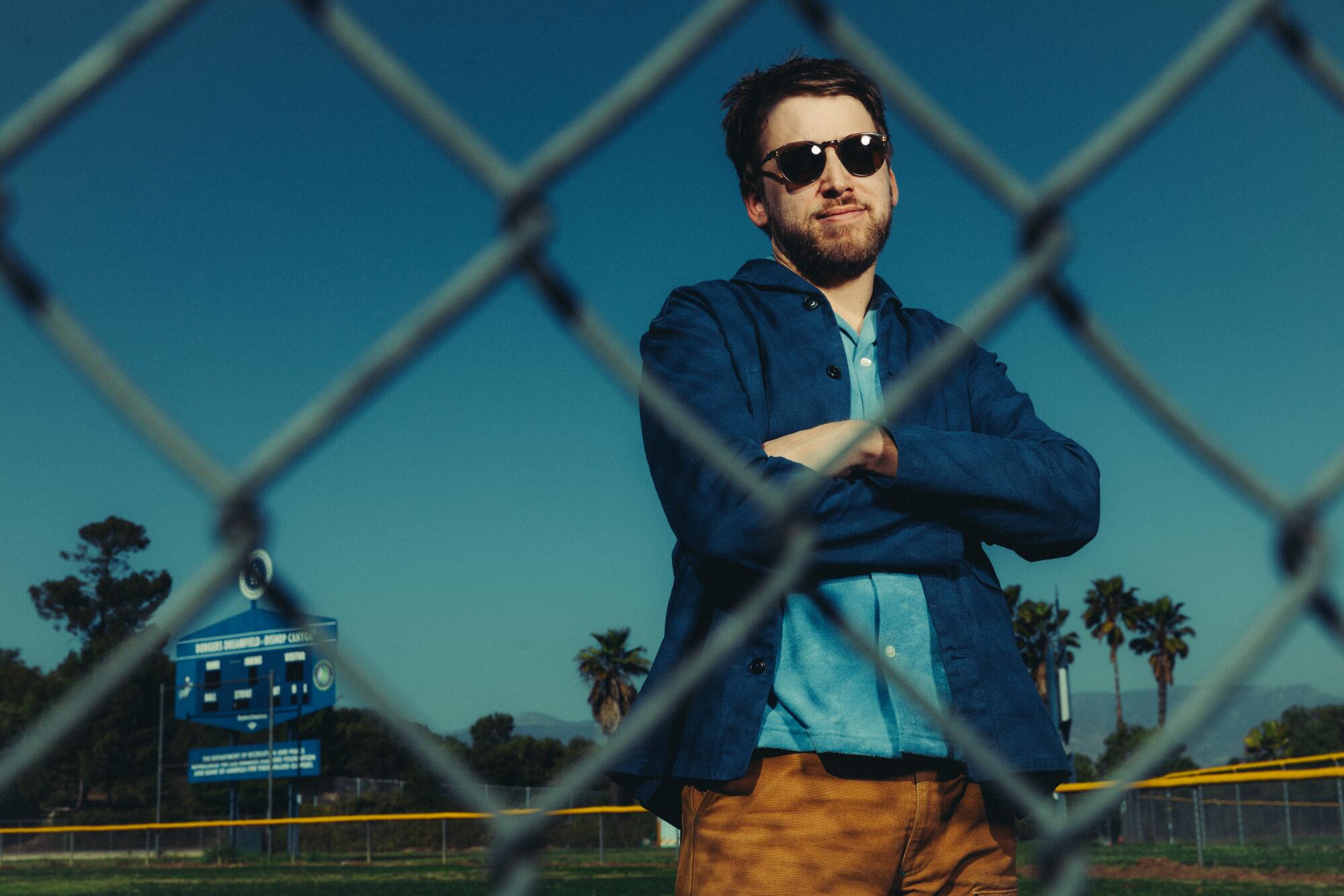 A man in shades stands on a baseball field behind a chain-link fence.