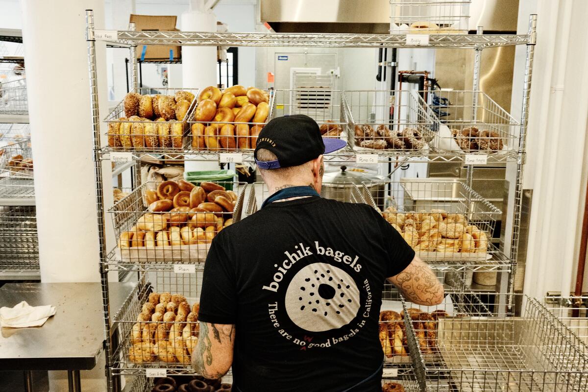 A person in a "Boichik Bagels" shirt, with back to camera, stands in front of racks of bagels