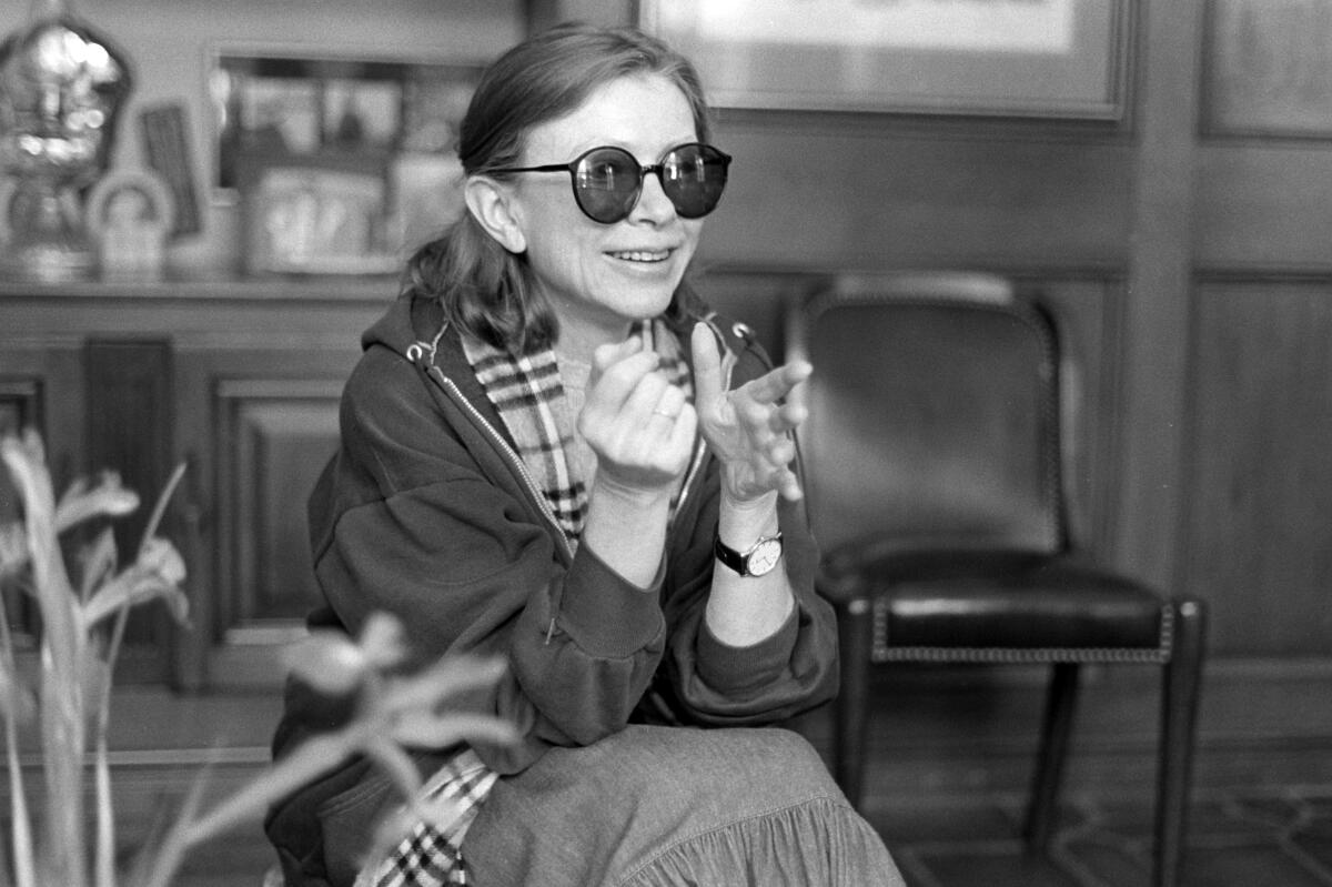 A photo of writer Joan Didion sitting down, wearing sunglasses and smiling at something off camera. 