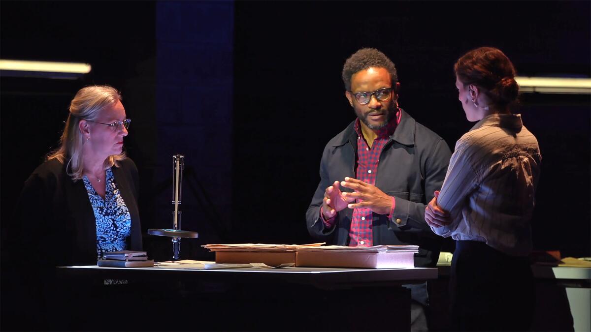 Standing next to a table, three people -- two female, one male -- have a serious conversation on stage.