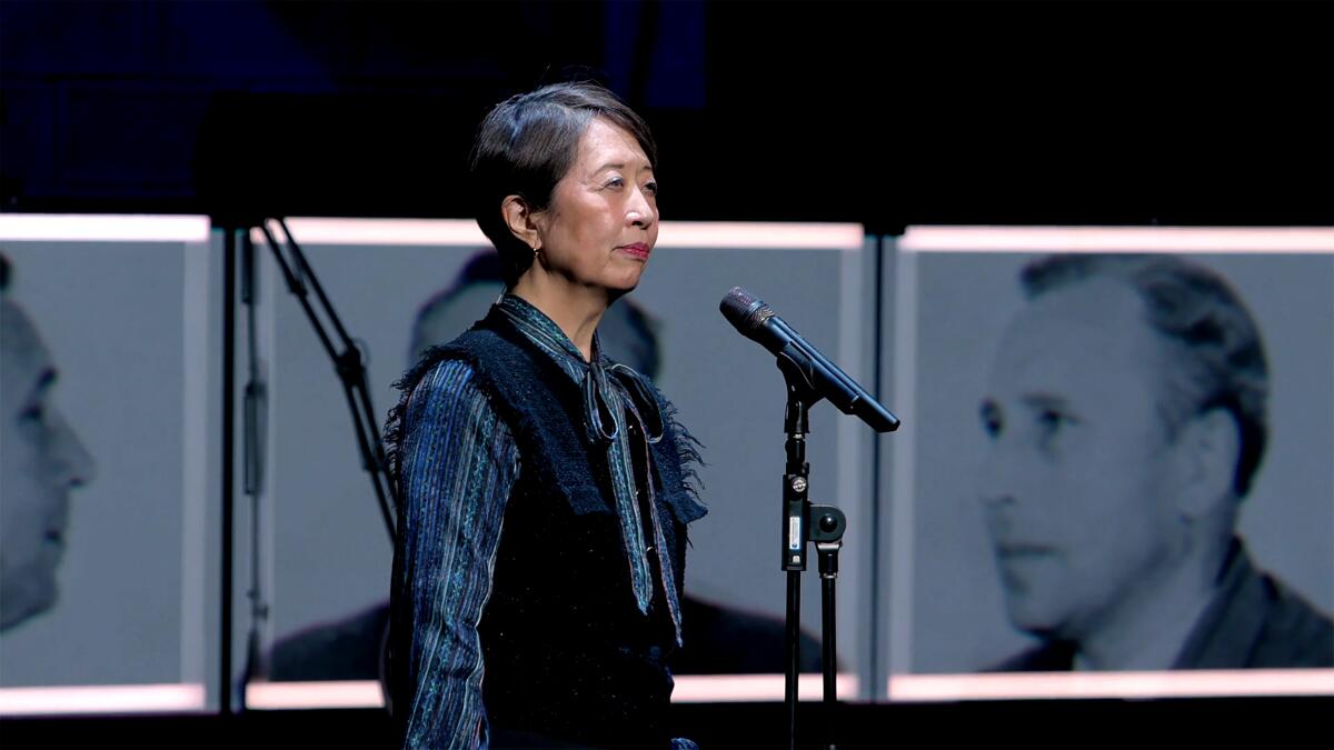 A woman stands behind a microphone stand, with black-and-white photos in the background, on stage.