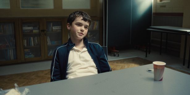 Owen Cooper in a white top and blue jacket sat at a table in a scene from Adolescence.