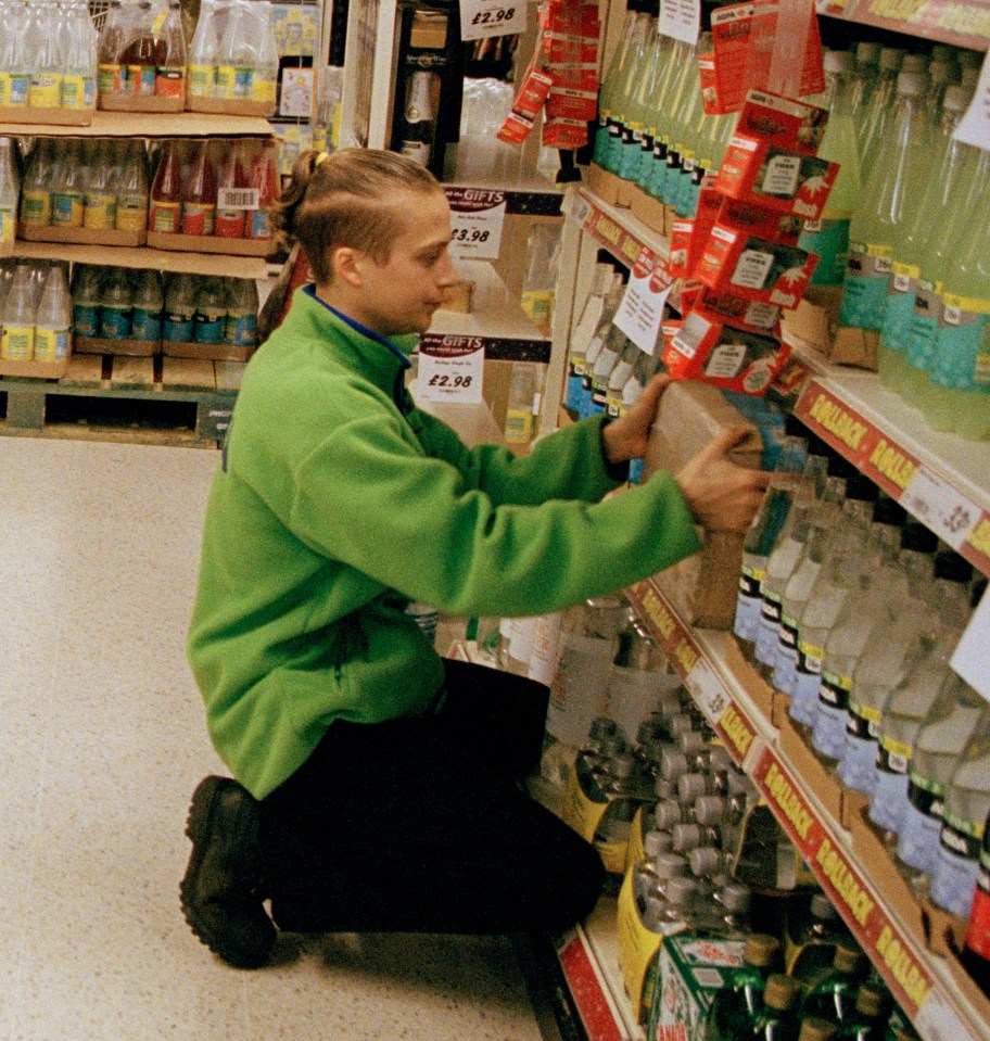 Woman shopping in a drinks aisle of a supermarket, with staff restocking shelves.