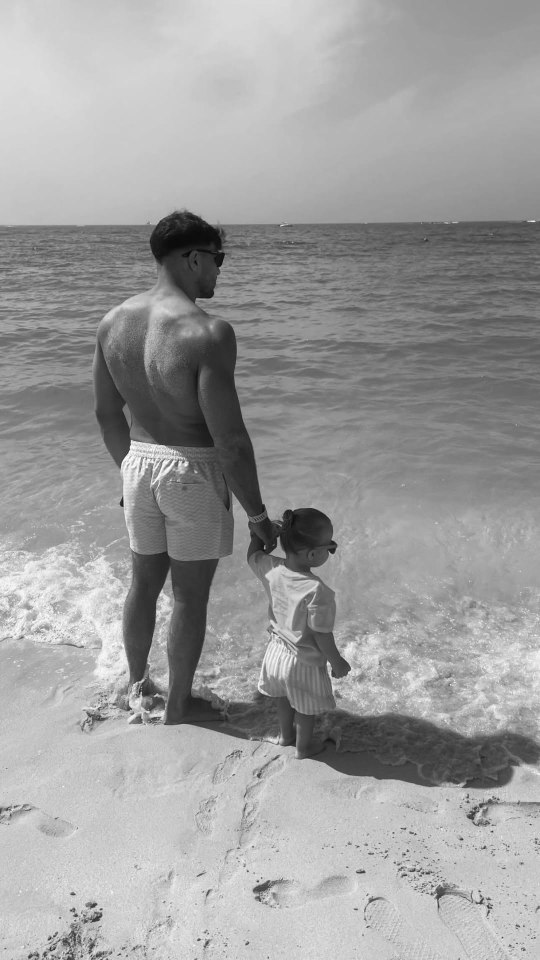 Black and white photo of a man and a toddler holding hands at the beach.