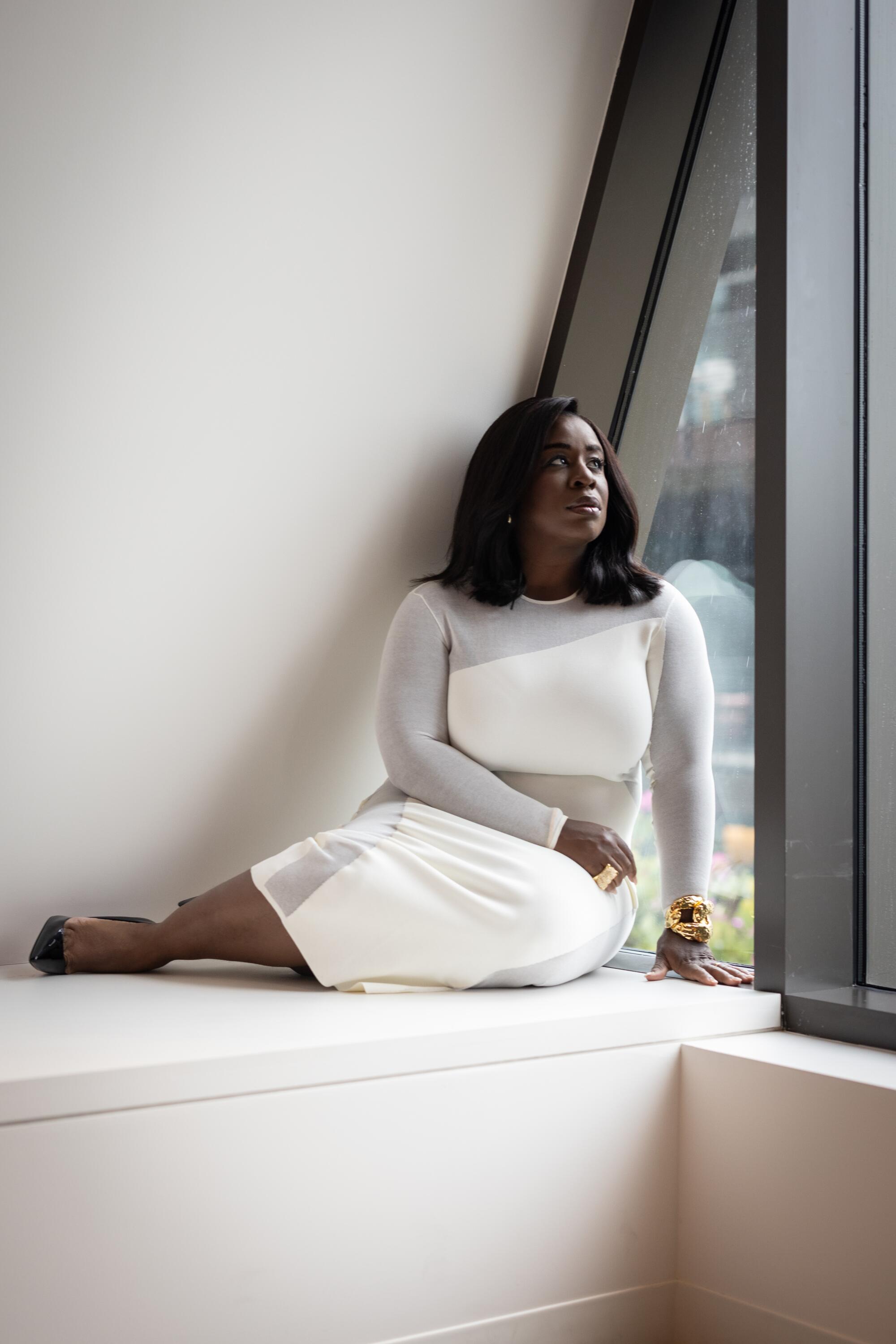 A woman in a white dress sits with her legs to the side near a window.