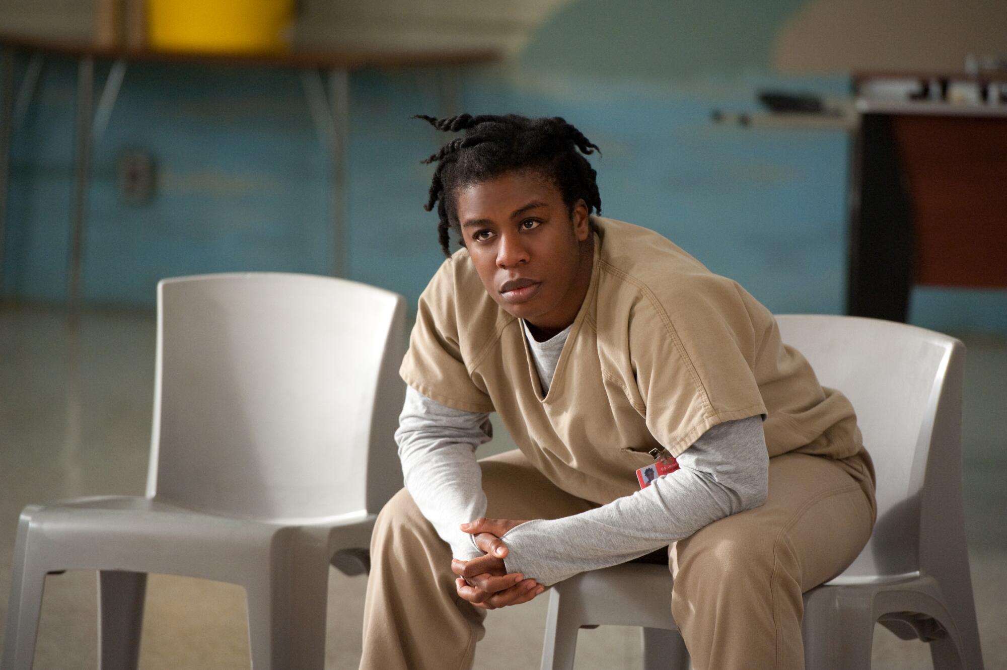 A woman in a tan prison jumpsuit with a gray long-sleeve shirt underneath sits leaning forward on a gray chair.