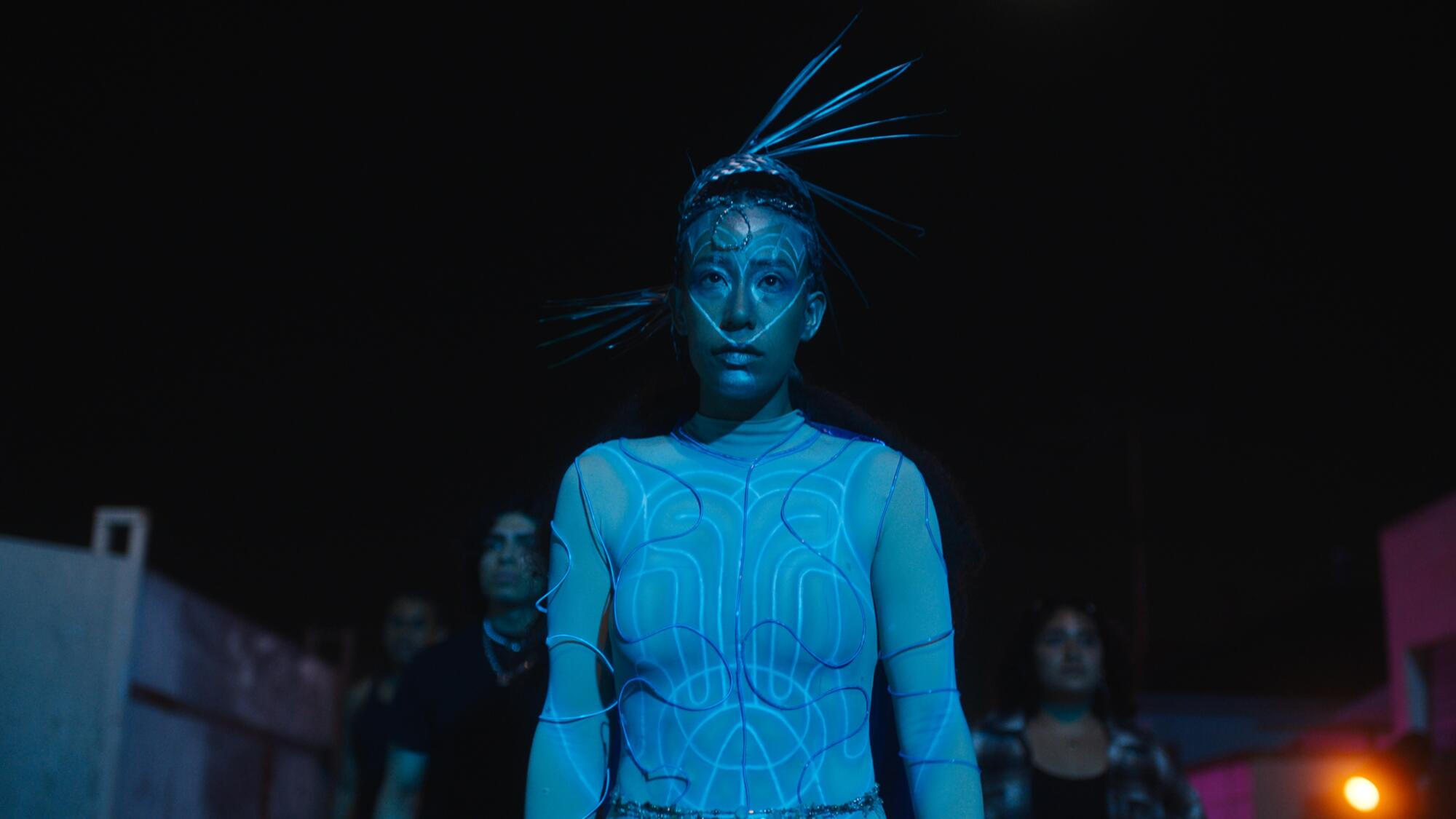 Artist Maria Maea is dressed as an alien in blue lighting.