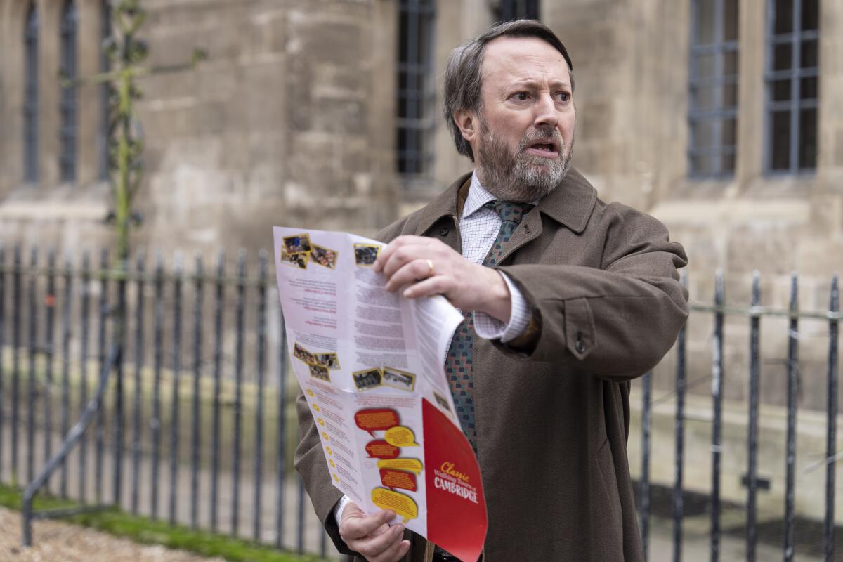 A man in a brown coat looks to the side as he's holding open a red, white and yellow pamphlet.