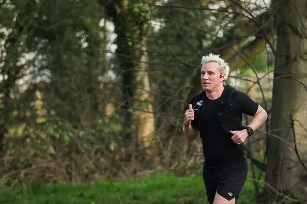 Jamie Laing in a black top and shorts running through a wooded area.