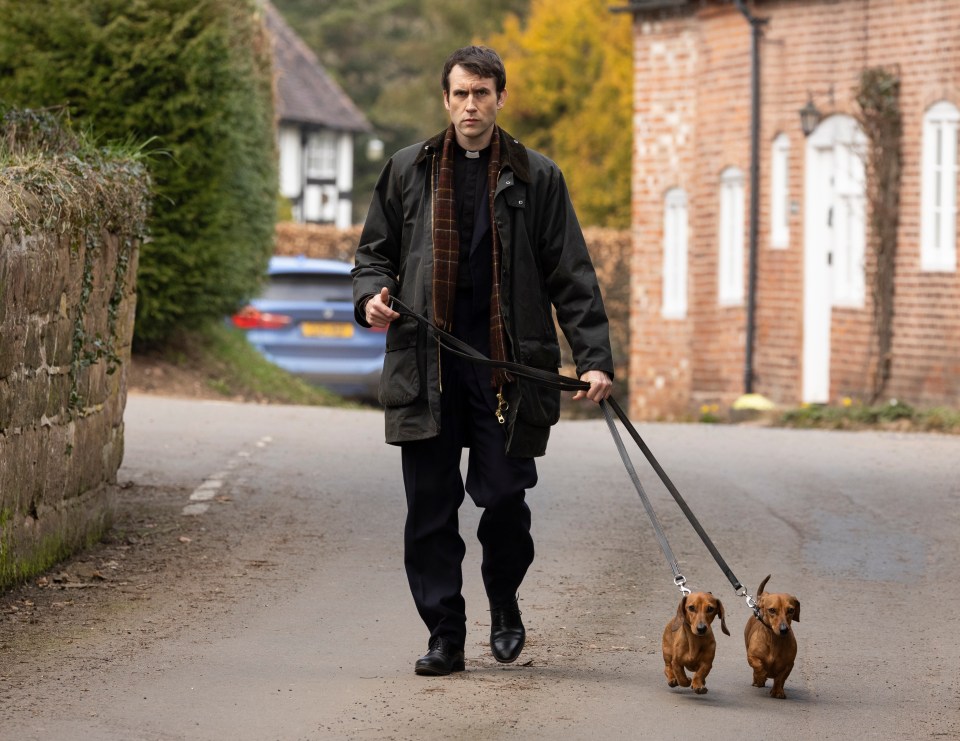 Matthew Lewis, actor known for playing Neville Longbottom in the Harry Potter films, walking two dachshunds on set of the TV drama *Murder Before Evensong*.