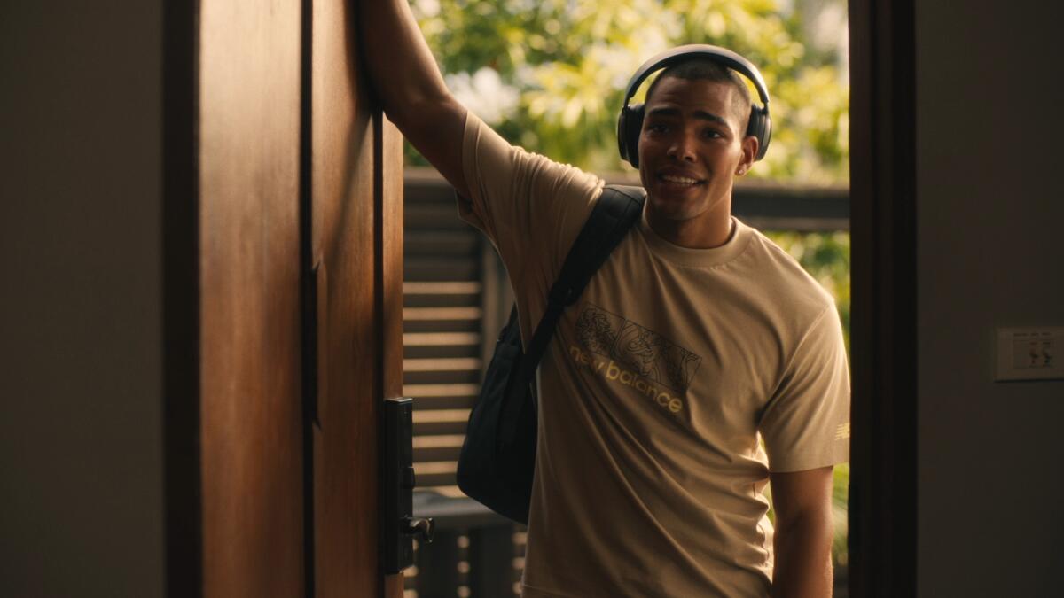 A man in a T-shirt wearing headphones leans against the frame of a doorway.