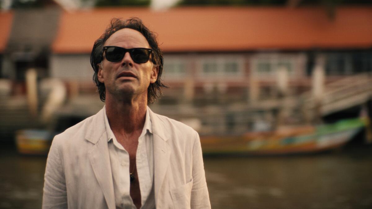 A man in a white shirt and suit wearing sunglasses looks upward. A river and a boat is seen behind him.