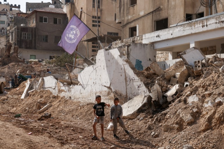 two children stand next to a destroyed building with a blue un flag flying from it