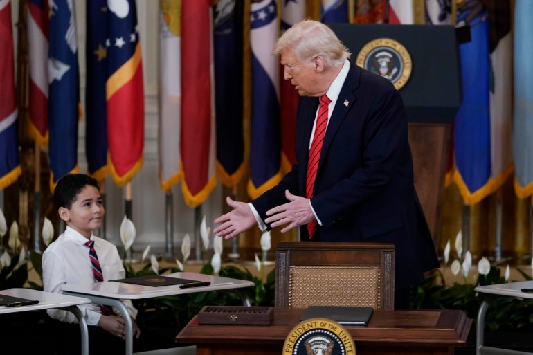 Donald Trump at a podium gestures to a kid seated next to him at a desk.