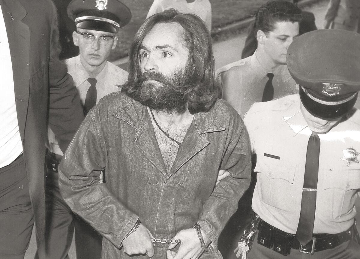 A black-and-white photo of a man with a dark beard and shoulder-length hair, in handcuffs, escorted by people in uniform