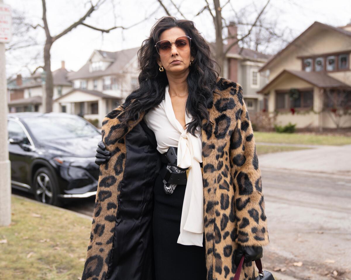 A woman in sunglasses and a leopard-print fur coat flashes a handgun in a holster on her hip.