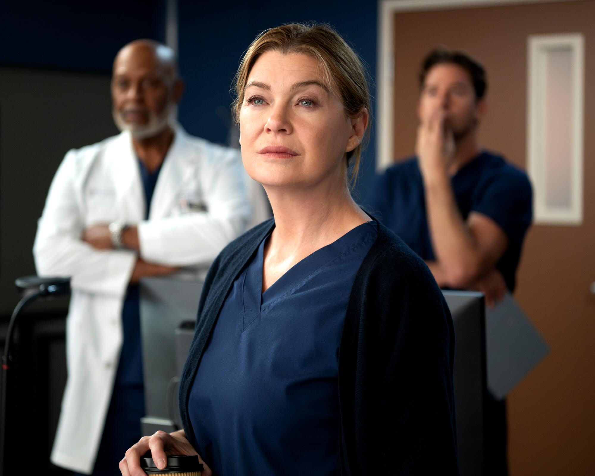 A woman in blue medical scrubs stares into the distance while two men stand behind her