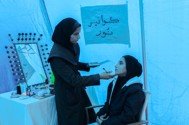 Noor pausing to check the makeup she has applied to Aya's face in the light filtering through the tent's blue plastic sidesåç