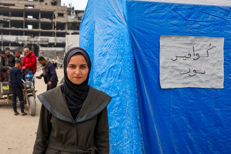 Noor stands in front of her tent with the handwritten sign on it. In the background is the destruction that is ubiquitous in Gaza today