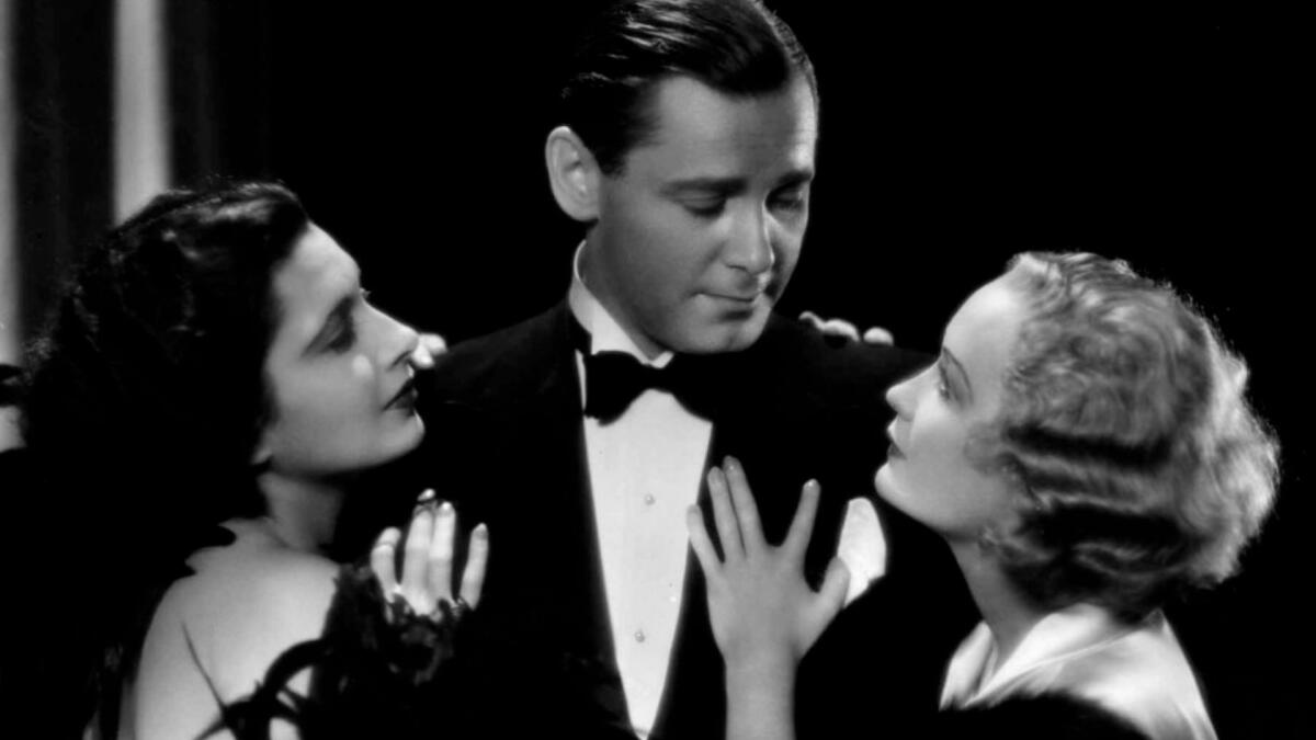 Two women embrace a man in a tuxedo.