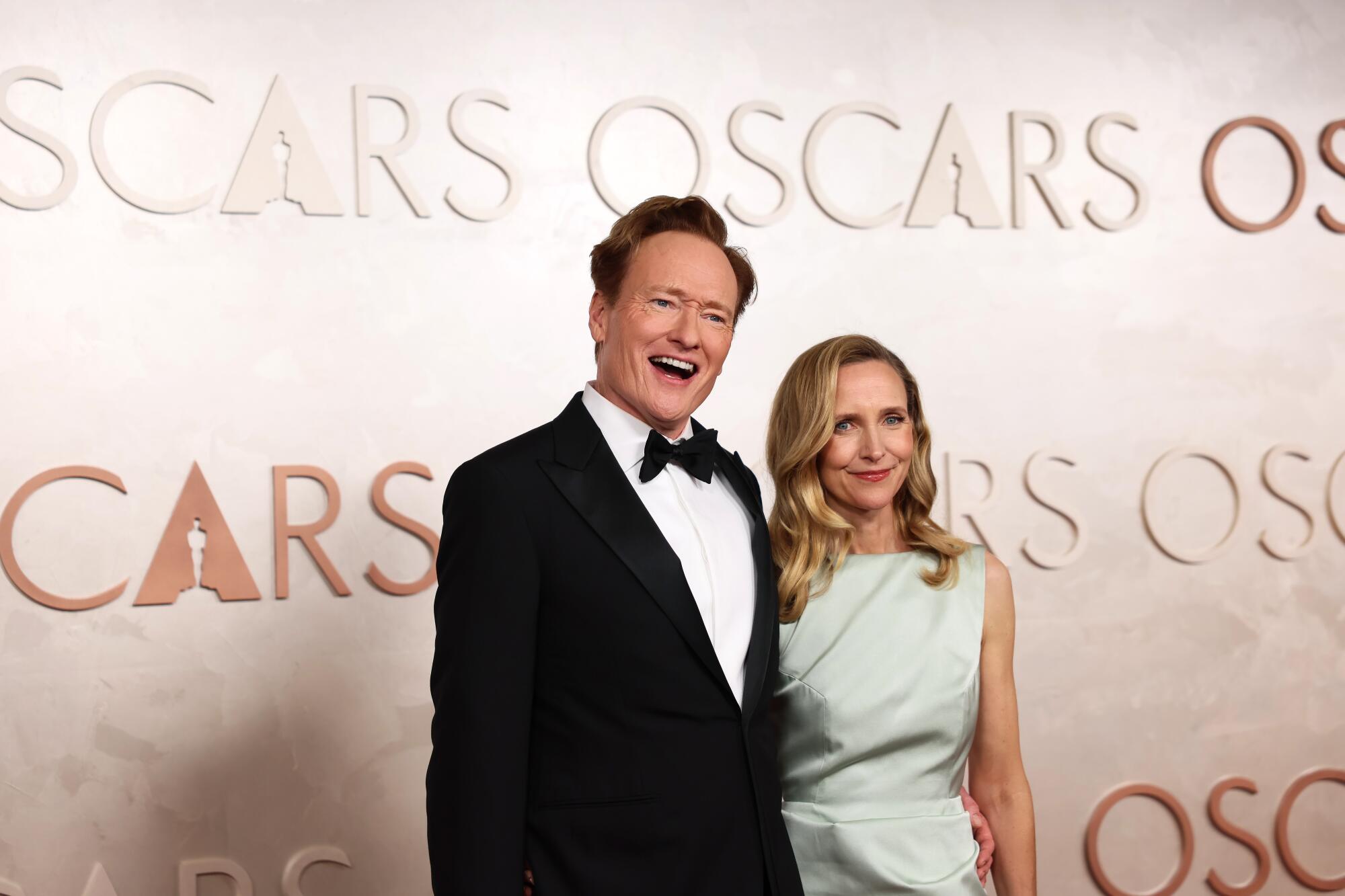 A man and a woman stand in front of a wall with the word "Oscars" repeated all over it.