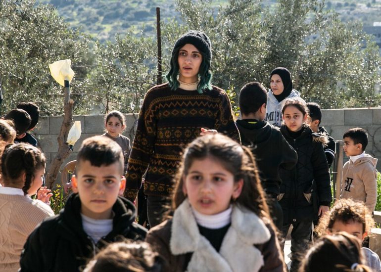 Shatha, in a woolly cap over her green hair, walks among a groups of children in the sunshine with olive trees in the background