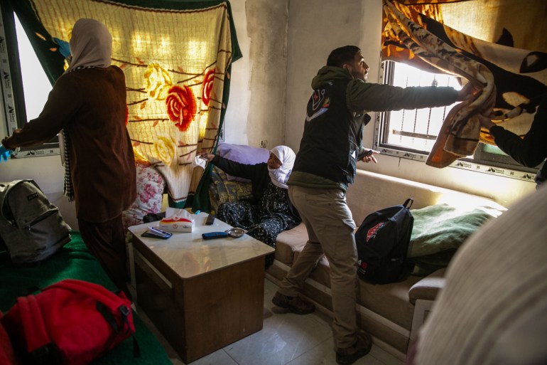 Two volunteers pull aside blankets stretched across the windows to air out a room for an elderly displaced woman who is seated on a sofa showing them what she needs them to do