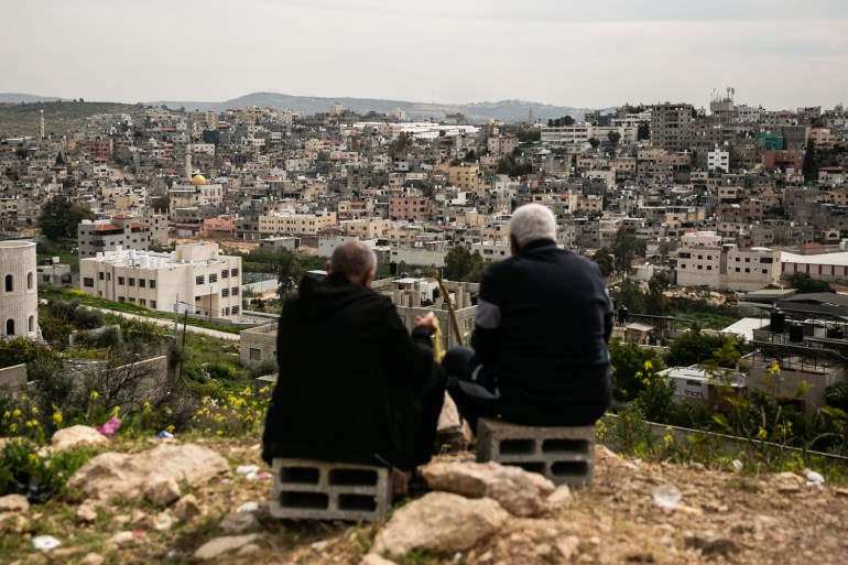 The backs of two elderly men, sitting on concrete breezeblocks, as the look down the hill towards where their homes used to be but are now out of reach