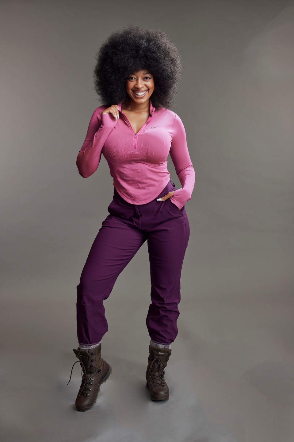 A woman smiling, wearing a pink top, purple pants, and brown boots.