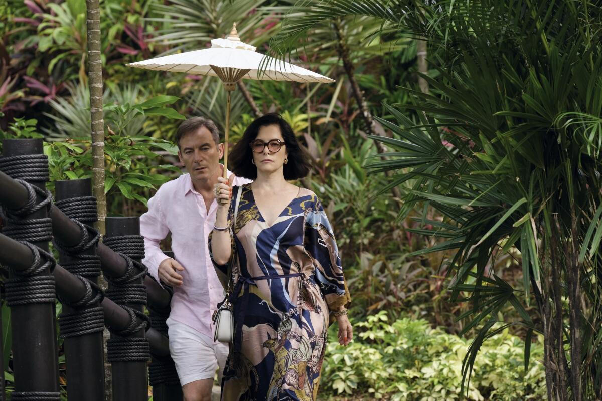 A man in a pink linen shirt and sorts walks behind a woman in a blue and pink dress holding a white umbrella.