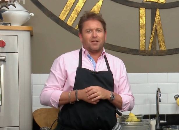 James Martin in a pink shirt and apron talking to the camera mid-show