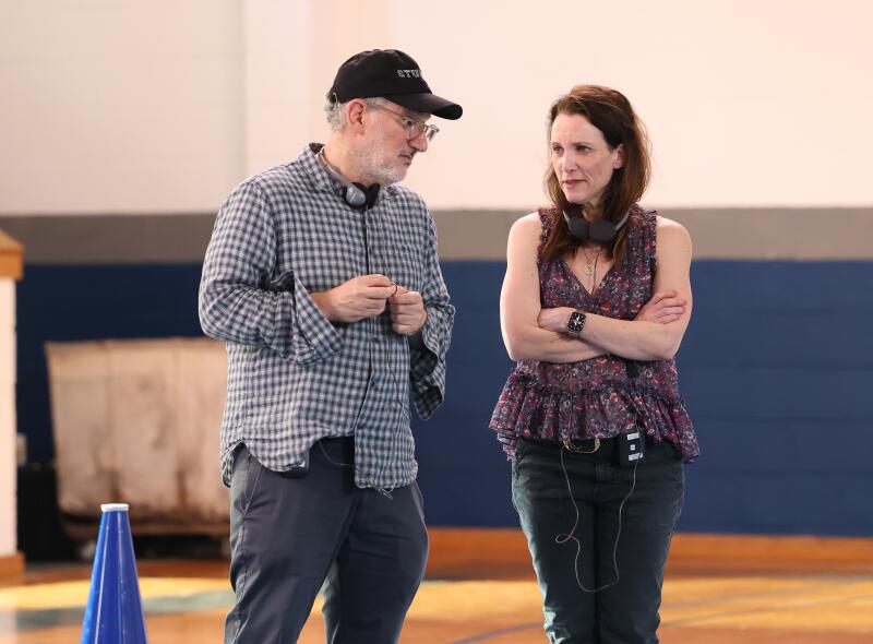 A man in a ball cap leans toward a woman in a floral sleeveless top in a gym.