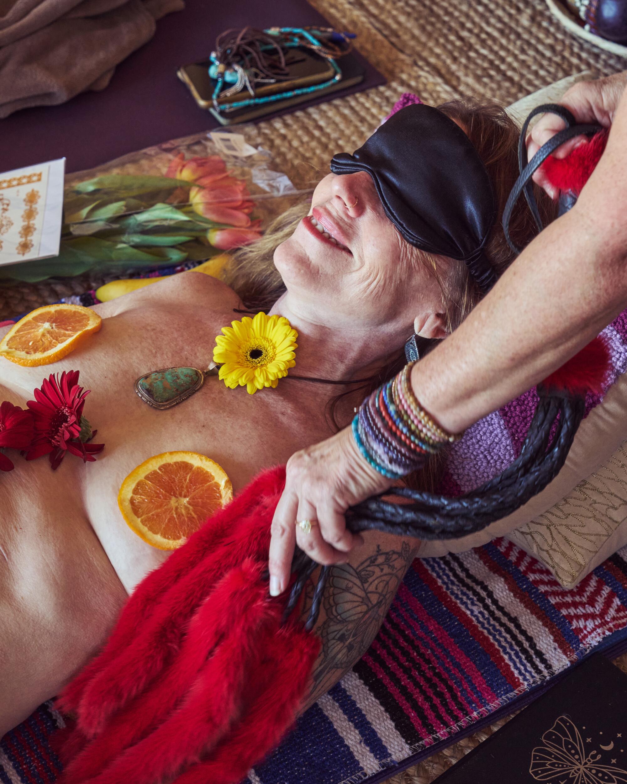 A woman reclines, blindfolded, with fruit and flowers on her body.