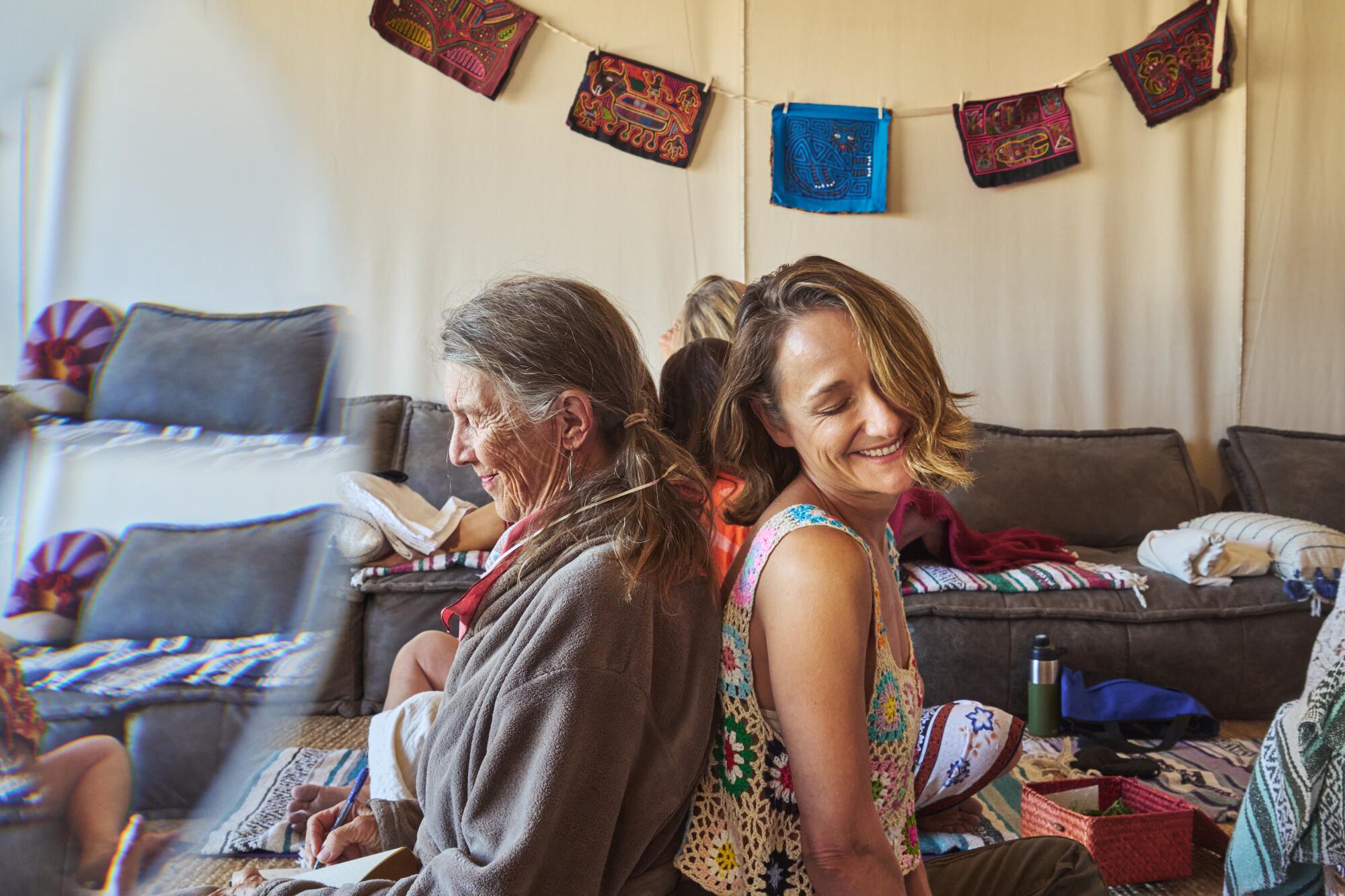 Two women sit back to back, smiling. 