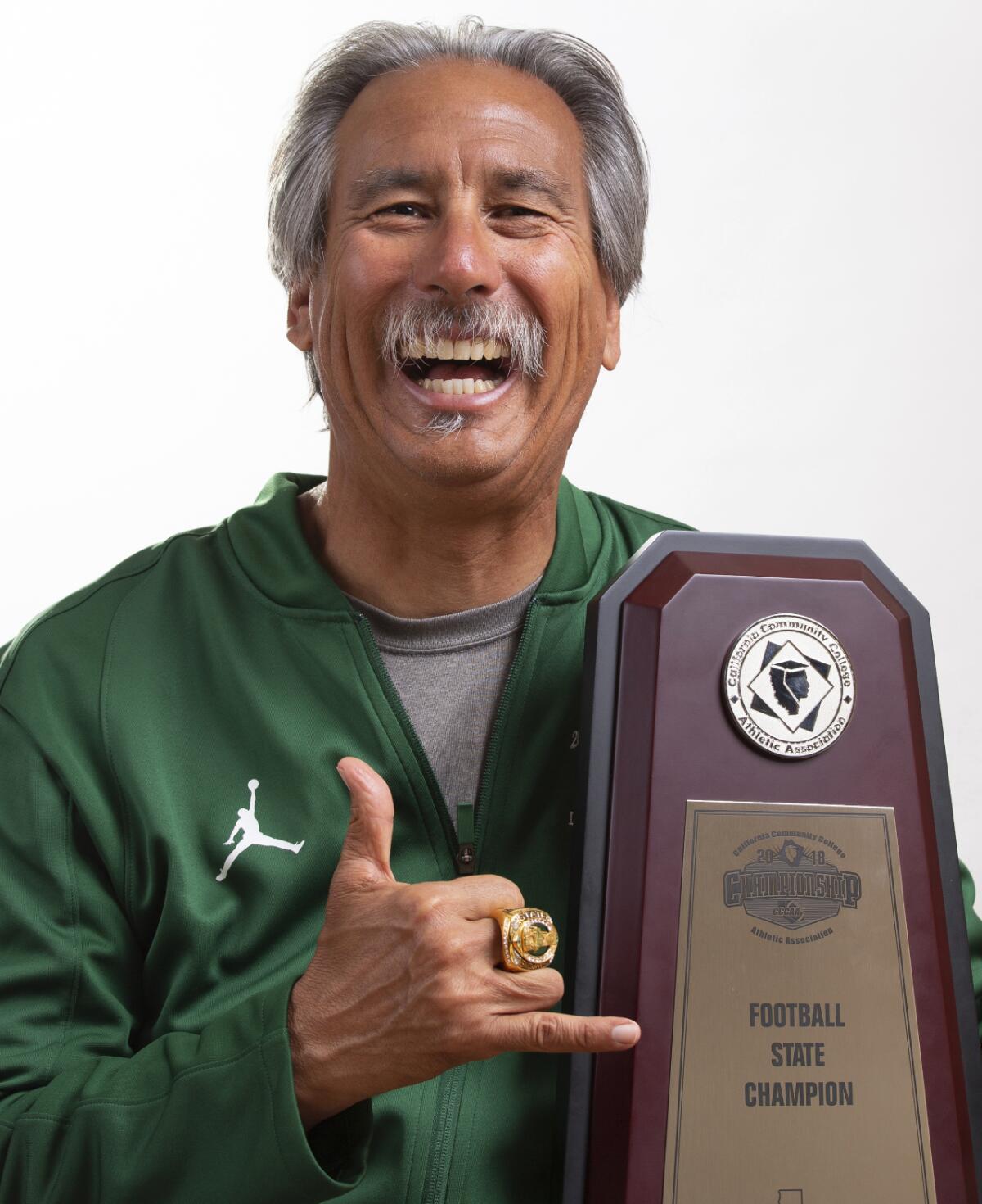 Laney College coaches John Beam smiles and gestures with his hand while holding the state championship trophy.