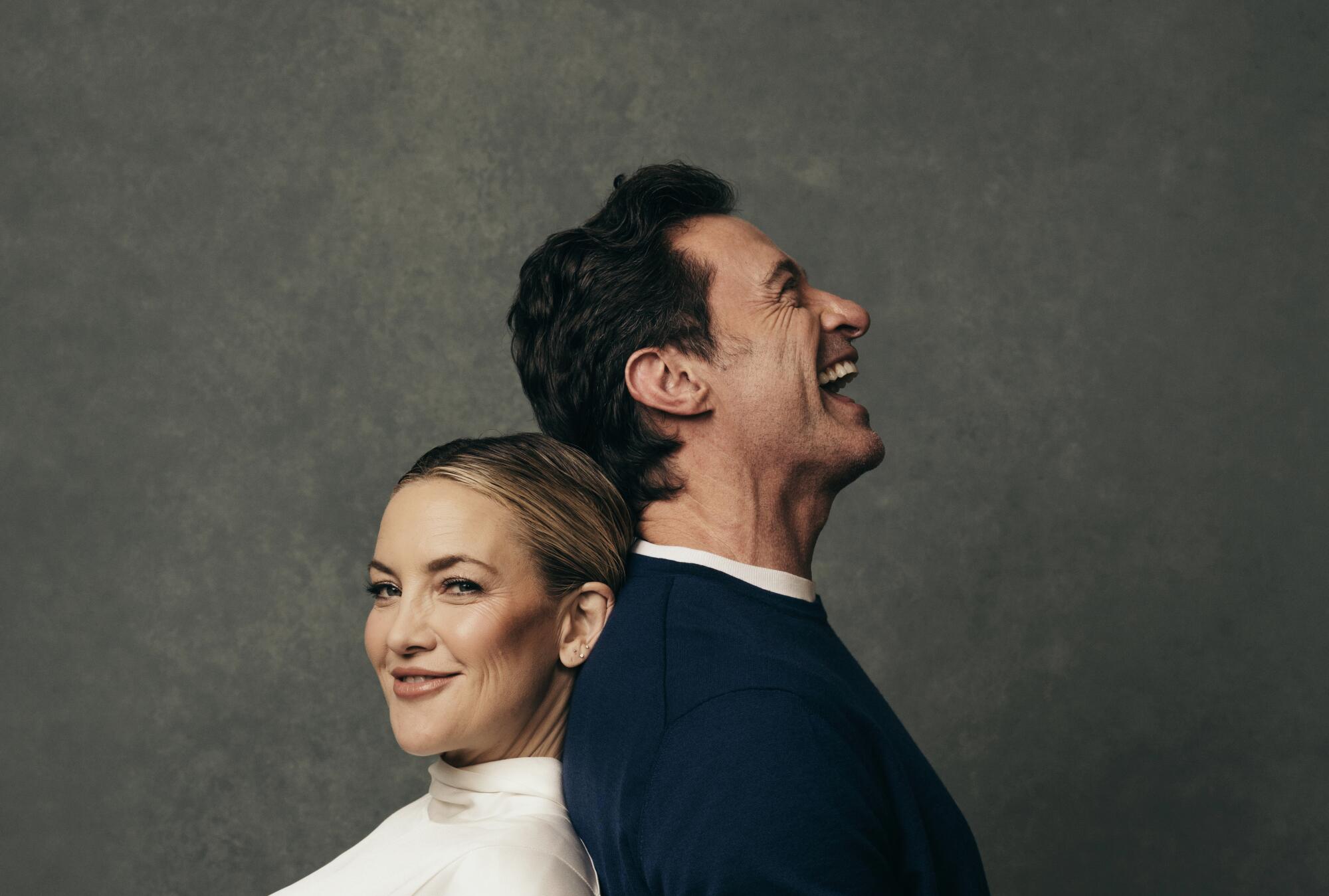 Two actors lean against each other, back to back, smiling.