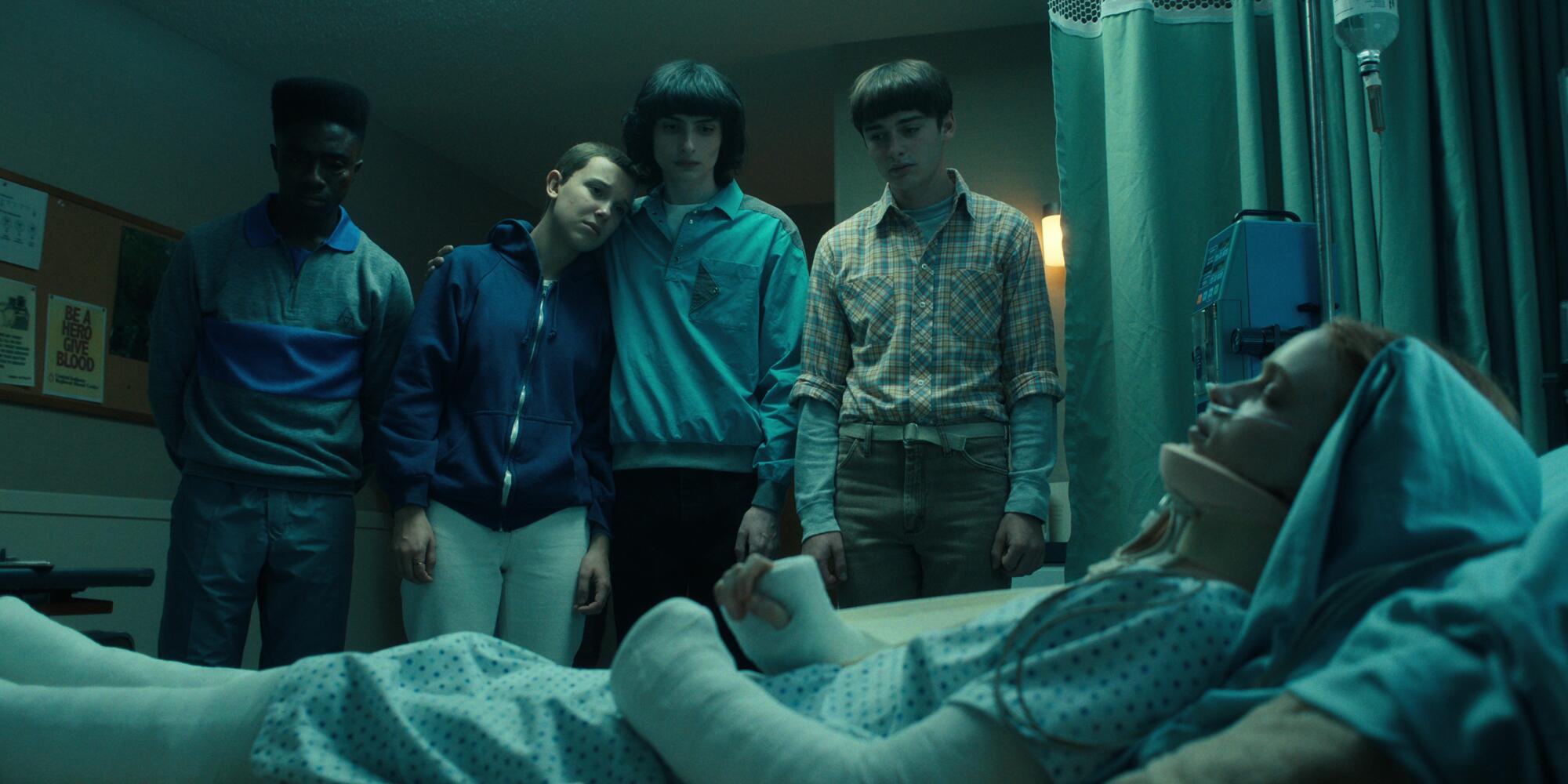 A still photo from "Stranger Things" of four people gathered around a girl in a hospital bed.