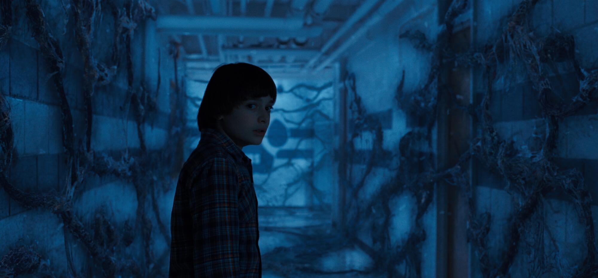 A still photo from Stranger Things of a boy in a dark hallway with vines on the walls