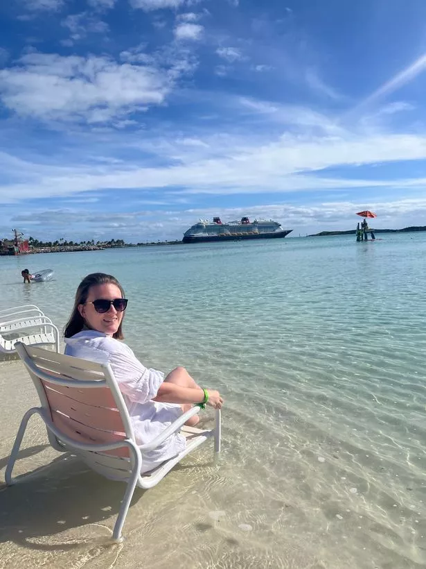 Julie sitting on a sunlounger by a beach with the Disney Destiny in the background