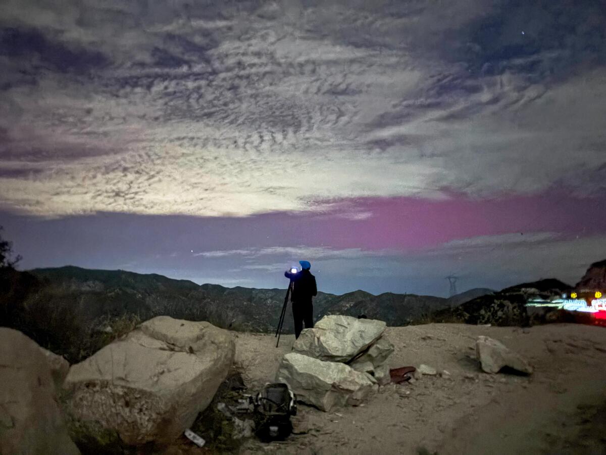 A silhouette of a person standing at a tripod and camera capturing a pinkish cloudy sky above a ridgeline.