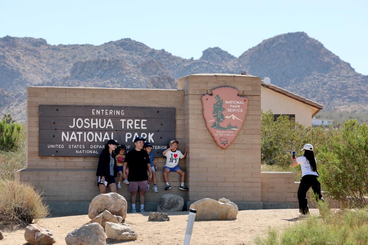 A family poses at the large tan brick Joshua Tree National Park sign near rocky hills.