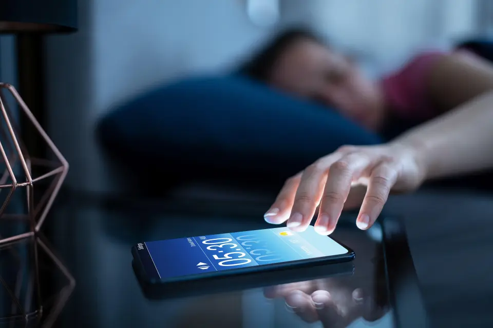 Woman's hand reaching for a smartphone displaying an alarm at 5:30 AM.