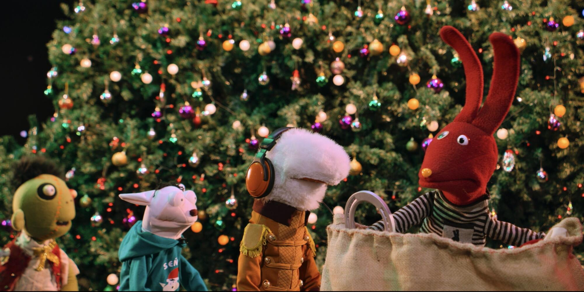 Four puppets standing next to each other in front of a giant Christmas tree decorated with colorful, shiny ornaments.