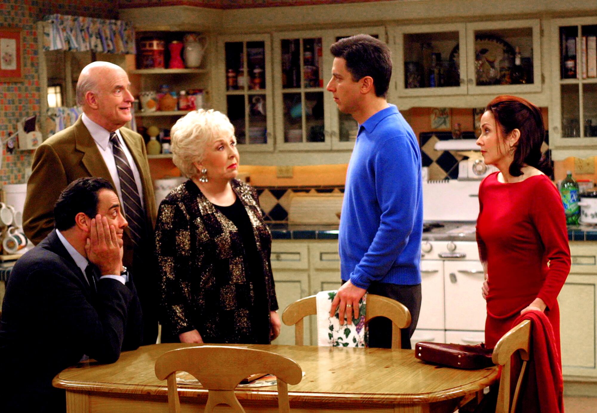 A older couple stands facing a younger couple as a man sits at a kitchen table and looks up at them.