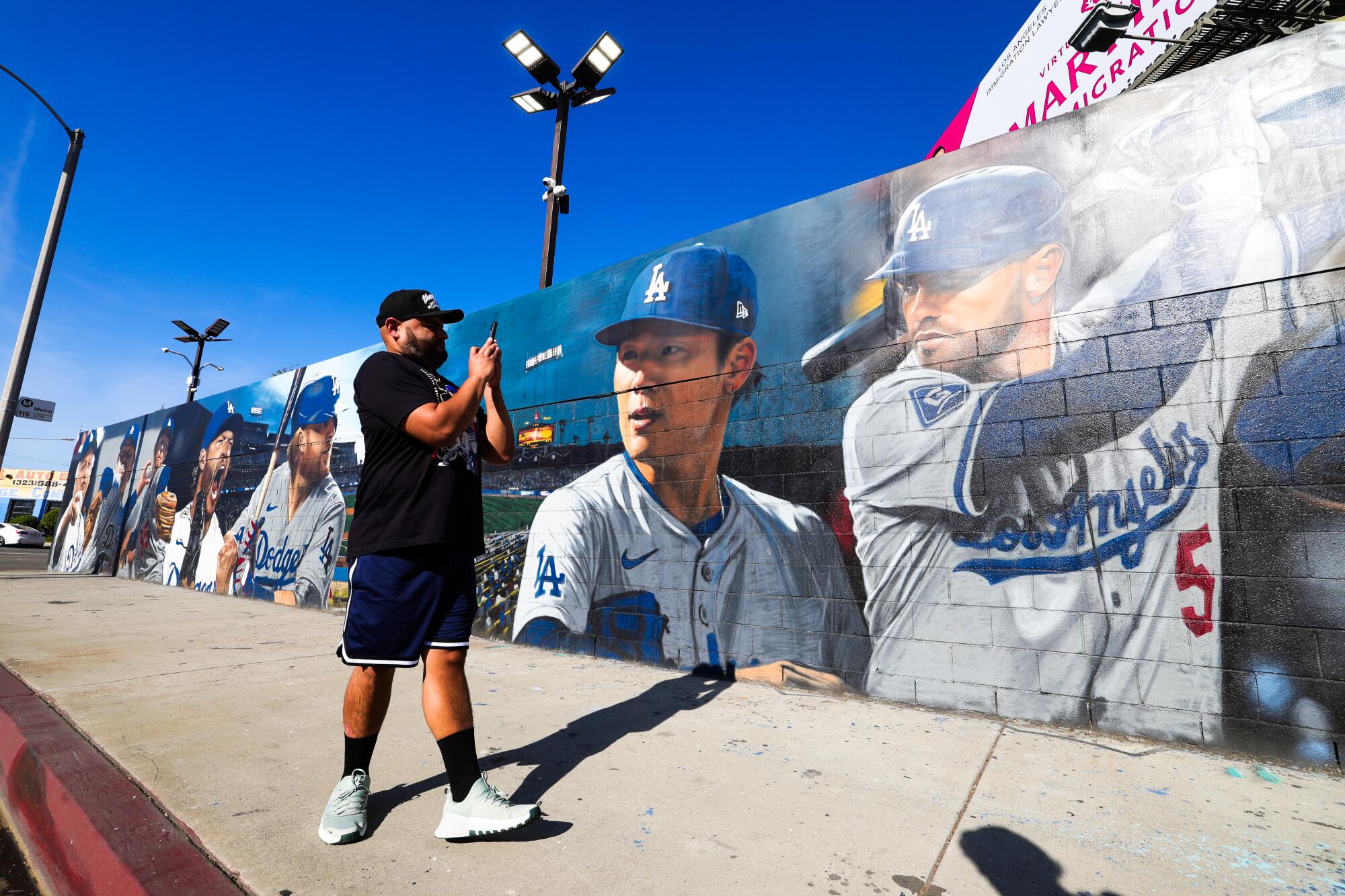 A man takes a picture of a sprawling Dodgers mural that features images of Freddie Freeman, Yoshinobu Yamamoto and others.