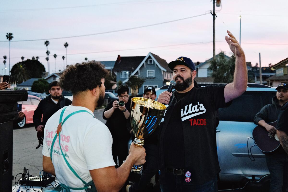 Victor Villa holds a large gold trophy in a parking lot at sundown. Memo Torres, right, presents Villa with the trophy.