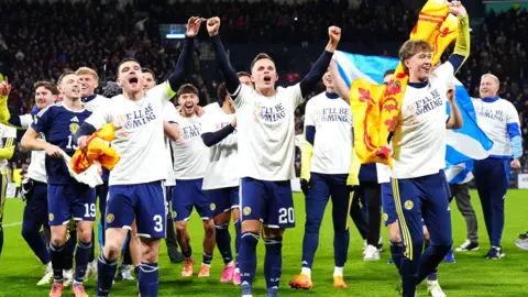 PA Media Football players celebrating on the pitch, wearing white shirts over their team kits and holding Scottish flags.